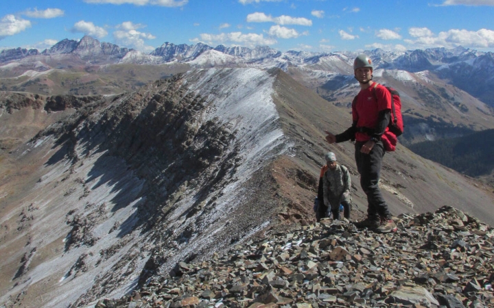 A person wearing safety gear stands atop a rocky summit in front of snow-capped mountains. Behind them, more people are making their way up to the summit. 